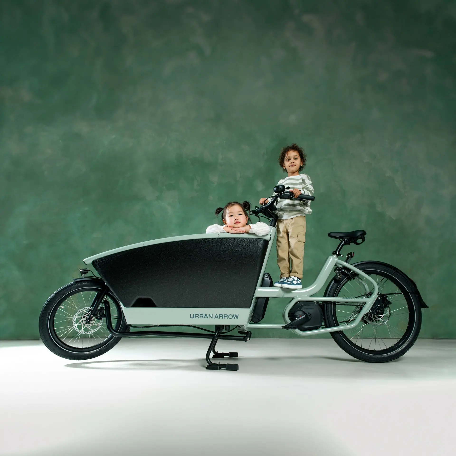 Zwei Kinder auf stehendem Lastenrad vor grüner Wand – spielerischer Moment und modernes Design vereint.