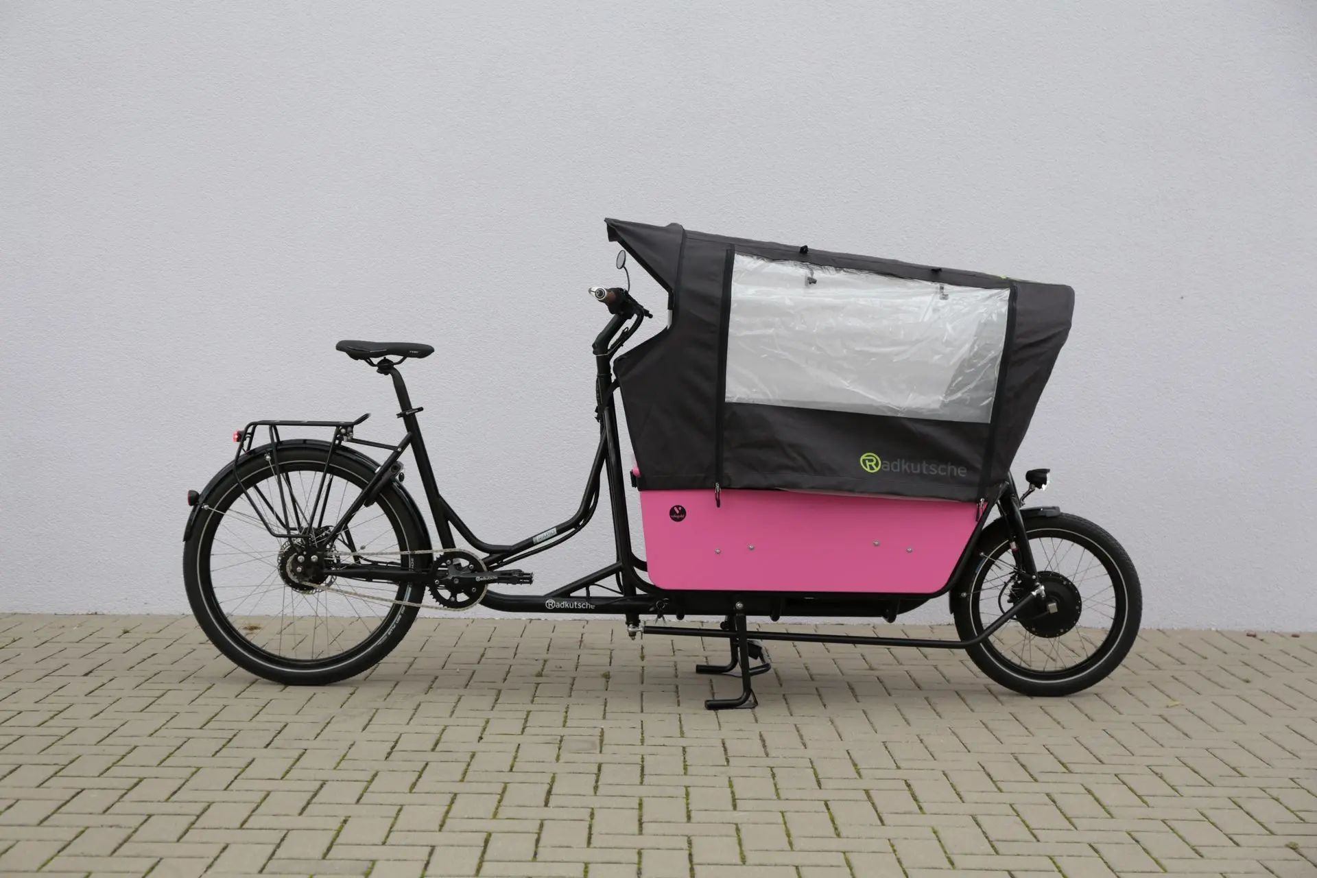 Radkutsche RAPID mit Transportwanne und Regenschutz-Zelt, abgestellt vor einer Hauswand – familienfreundliche Ausstattung.