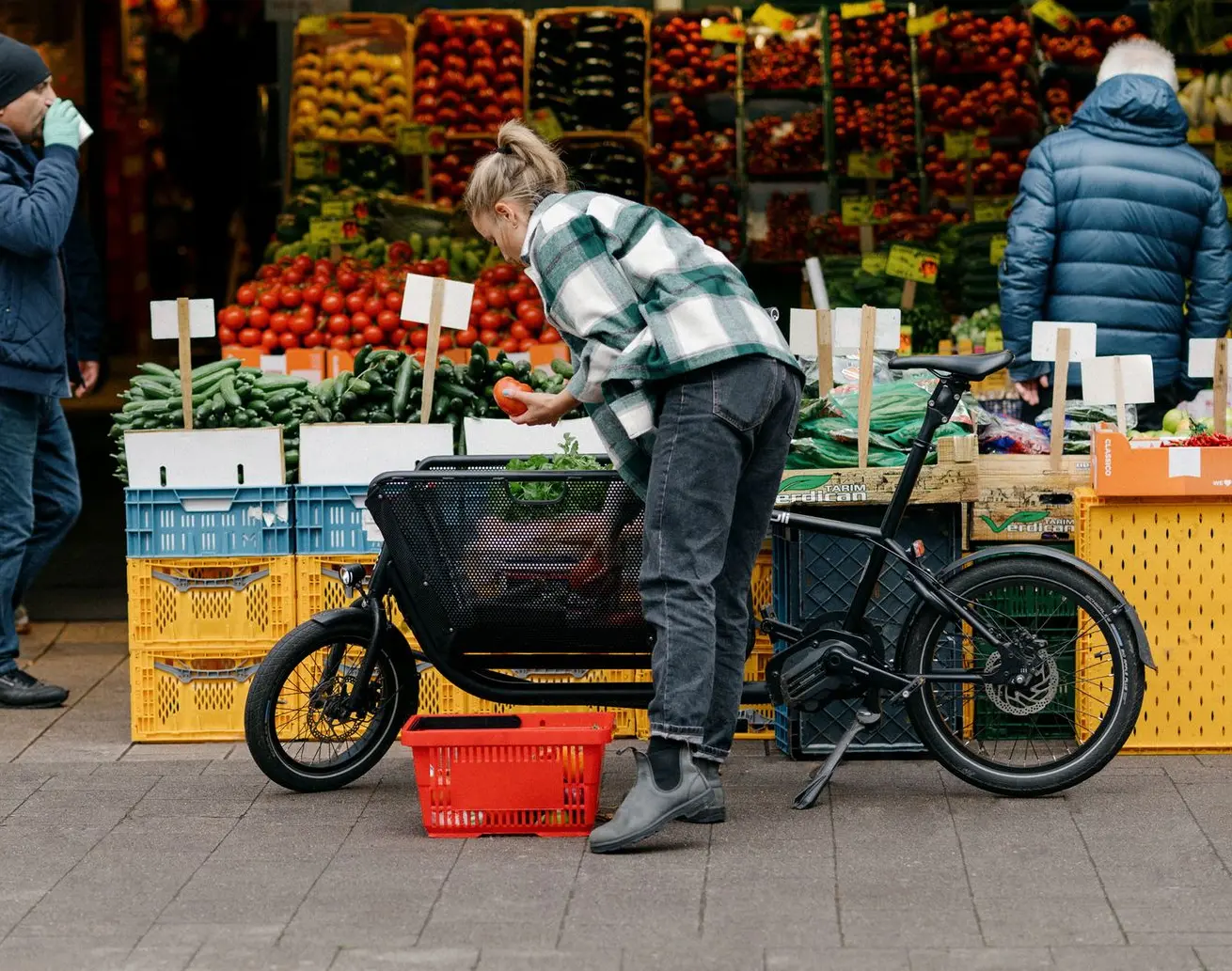 MULI Lastenrad beim Einkauf: kompakt, praktisch und mitten im Stadtleben. Person lädt Einkauf in ein MULI Lastenrad, im Hintergrund Marktstand mit frischem Gemüse.